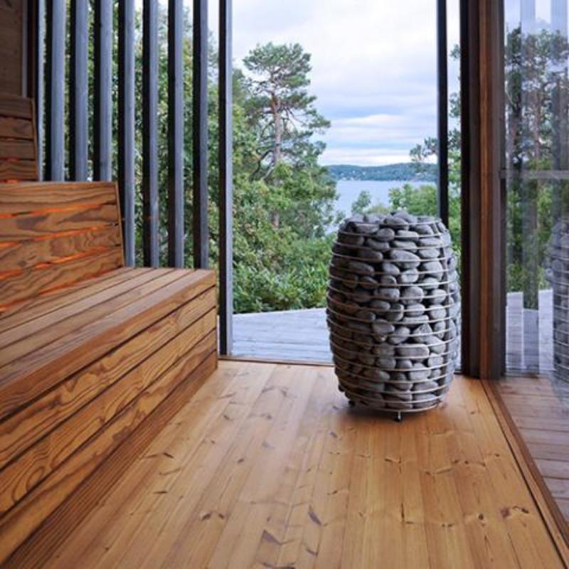 12 kW Electric Sauna Heater - the sauna heater sitting on the floor of a sauna with 2 perpendicular glass walls and a view of a lake in the distance and trees