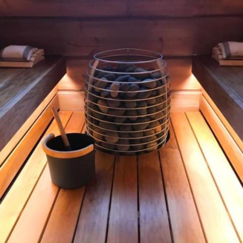 HIVE Series 18 kW Sauna Heater - sauna heater installed in the floor of the sauna with a bucket and ladle beside it. Backlighting from under the benches.