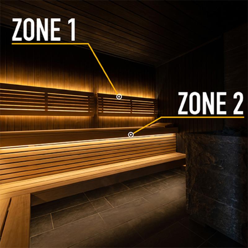 A wooden sauna interior with two labeled zones, Zone 1 and Zone 2, pointing to warm ambient lighting strips mounted on the wooden walls and benches. The room has dark wood paneling, light wood slat benches, dark stone flooring, and a dark stone vessel at the right edge.