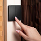 A close-up of a person's hand with a French manicure pressing a black two-switch wall dimmer or light switch mounted on a light wood door frame.