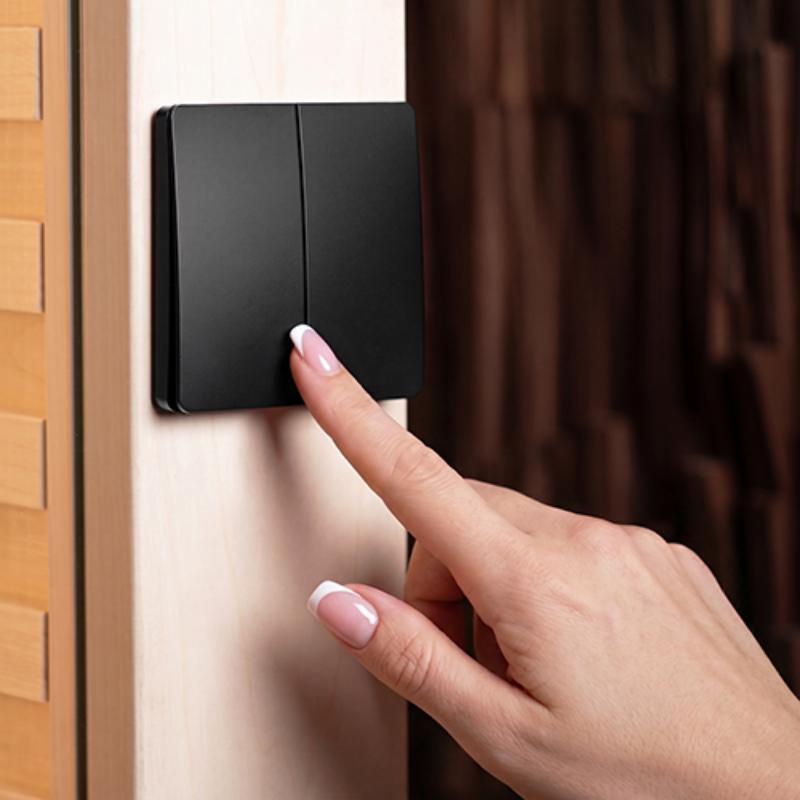 A close-up of a person's hand with a French manicure pressing a black two-switch wall dimmer or light switch mounted on a light wood door frame.